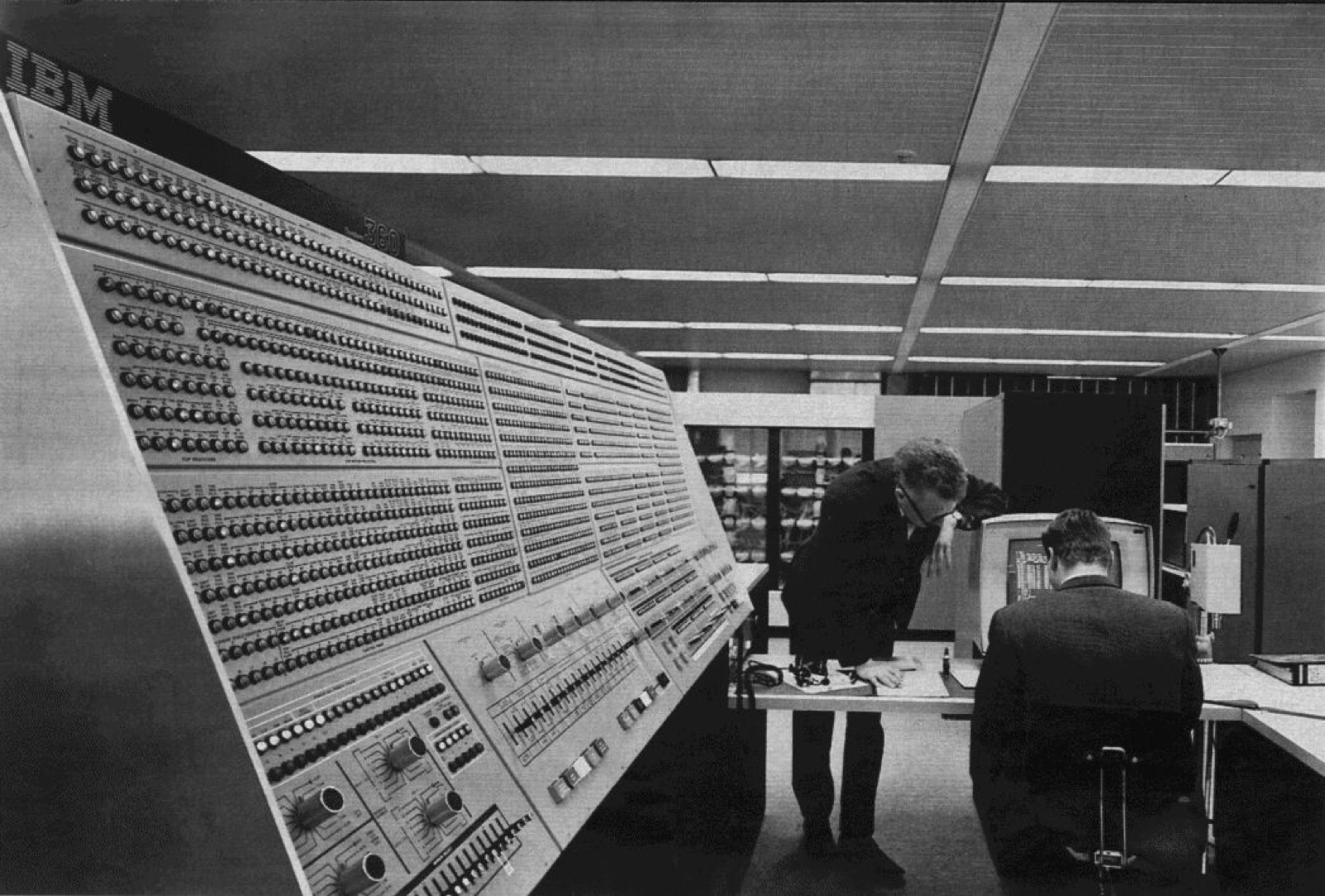 The switch-and-light-covered operator console of an IBM System/360 Model 91 mainframe, with two operators working at a desk behind it, late 1960s.