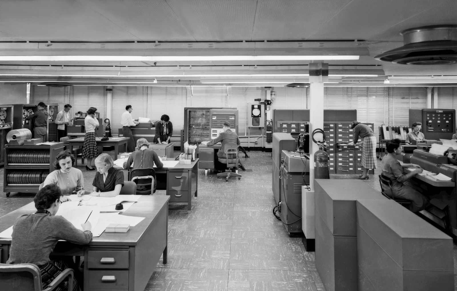 A 1957 NACA computing room: staff at desks working with paper and tape machines alongside an IBM 704 mainframe and its tape drives.