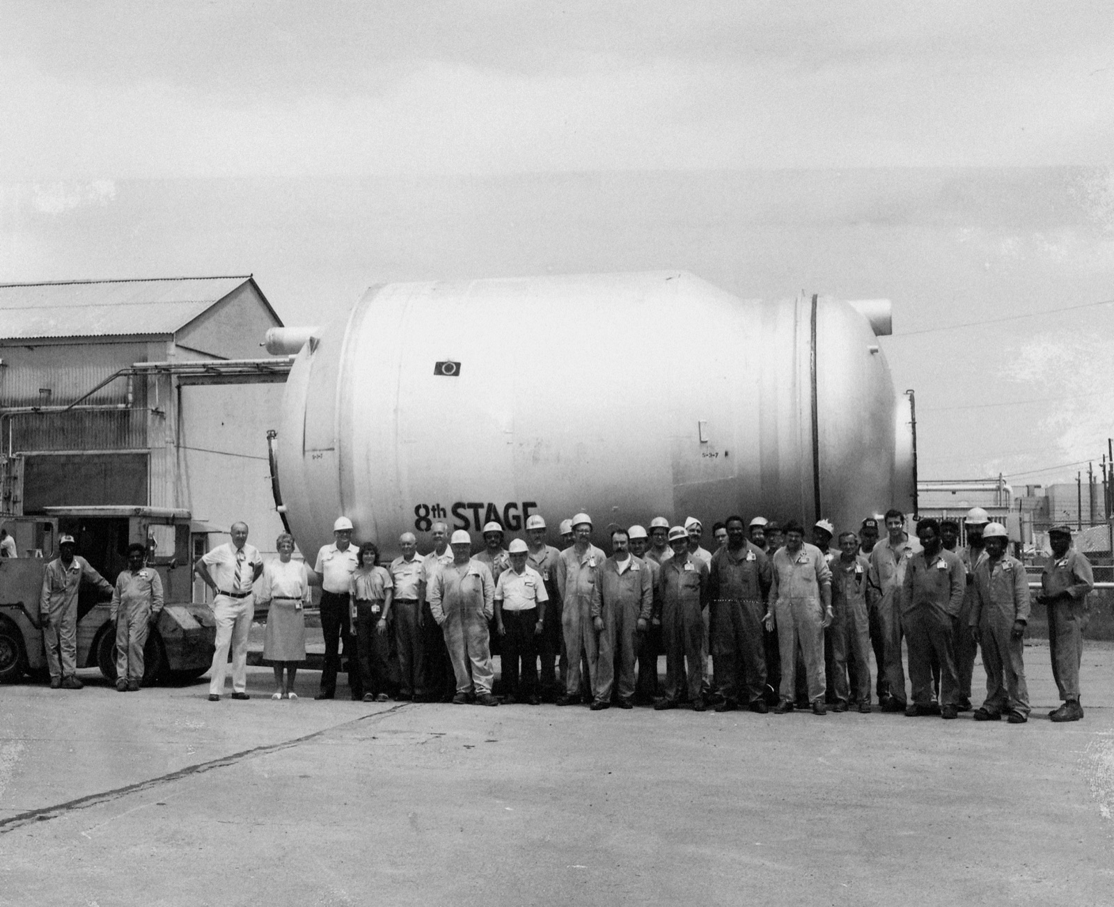 Workers in hard hats and coveralls posing outdoors in front of a huge cylindrical uranium-enrichment converter marked 8th Stage at the Paducah Gaseous Diffusion Plant in Kentucky.