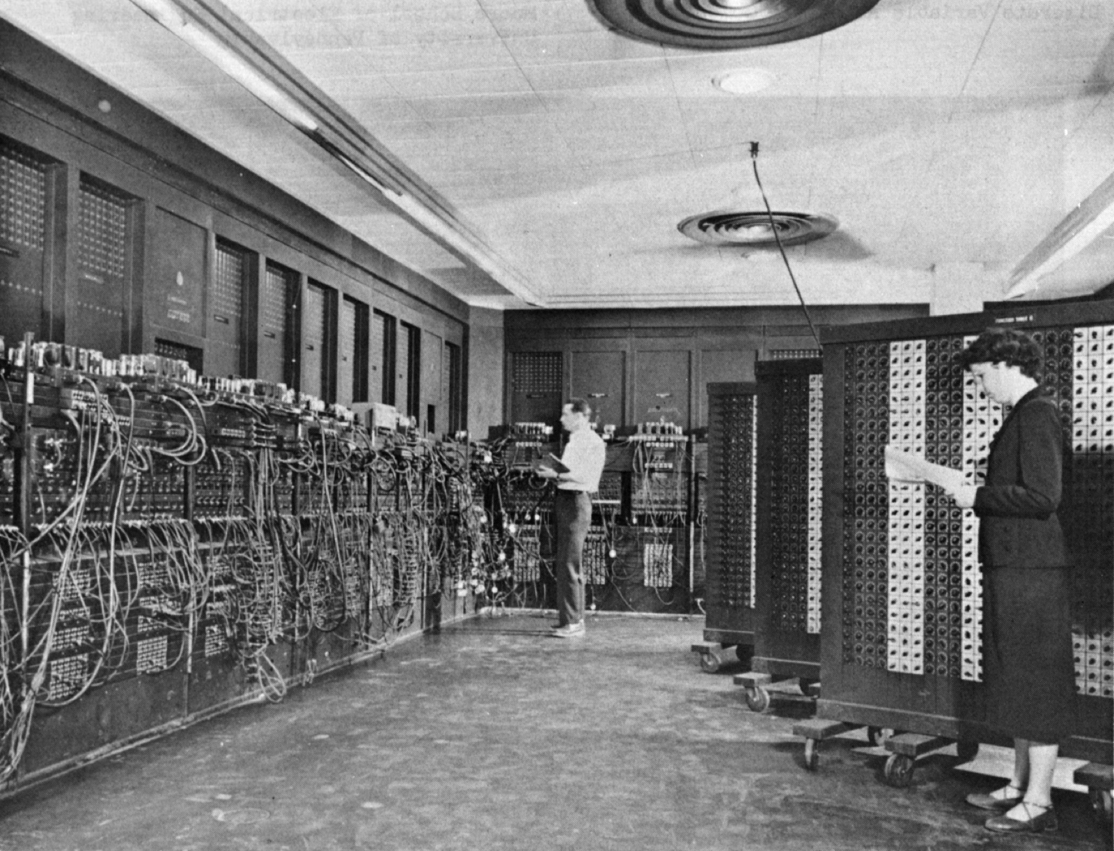 A black-and-white photograph of two people, Glen Beck and Betty Snyder, standing at the ENIAC in building 328 of the Army Ballistic Research Laboratory, setting the cable-strewn panels and portable function tables of the room-sized machine.