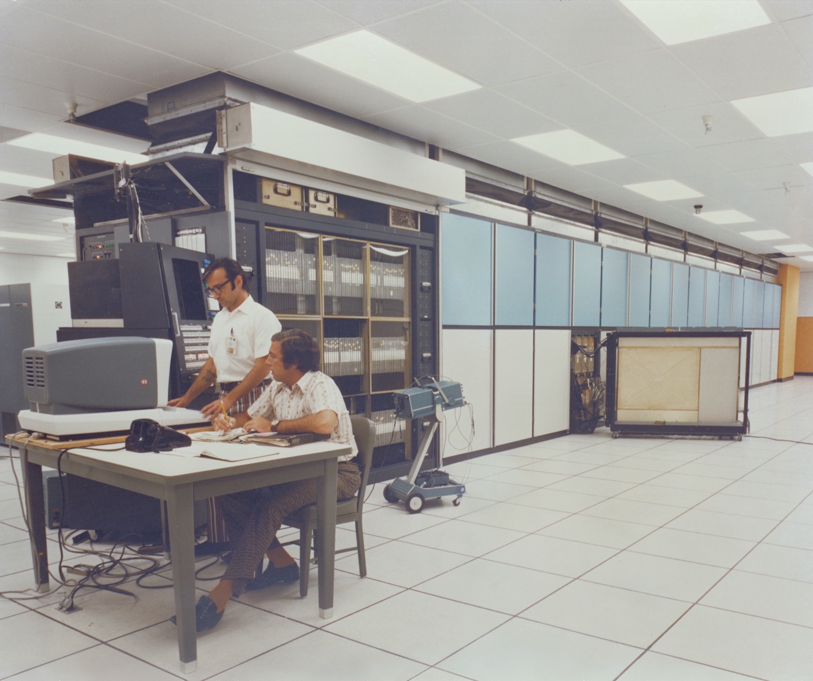 Two technicians, V. Tosti standing and S. Kravity seated at a work table, in front of the long cabinet row of the ILLIAC IV parallel computer at NASA Ames in 1972.