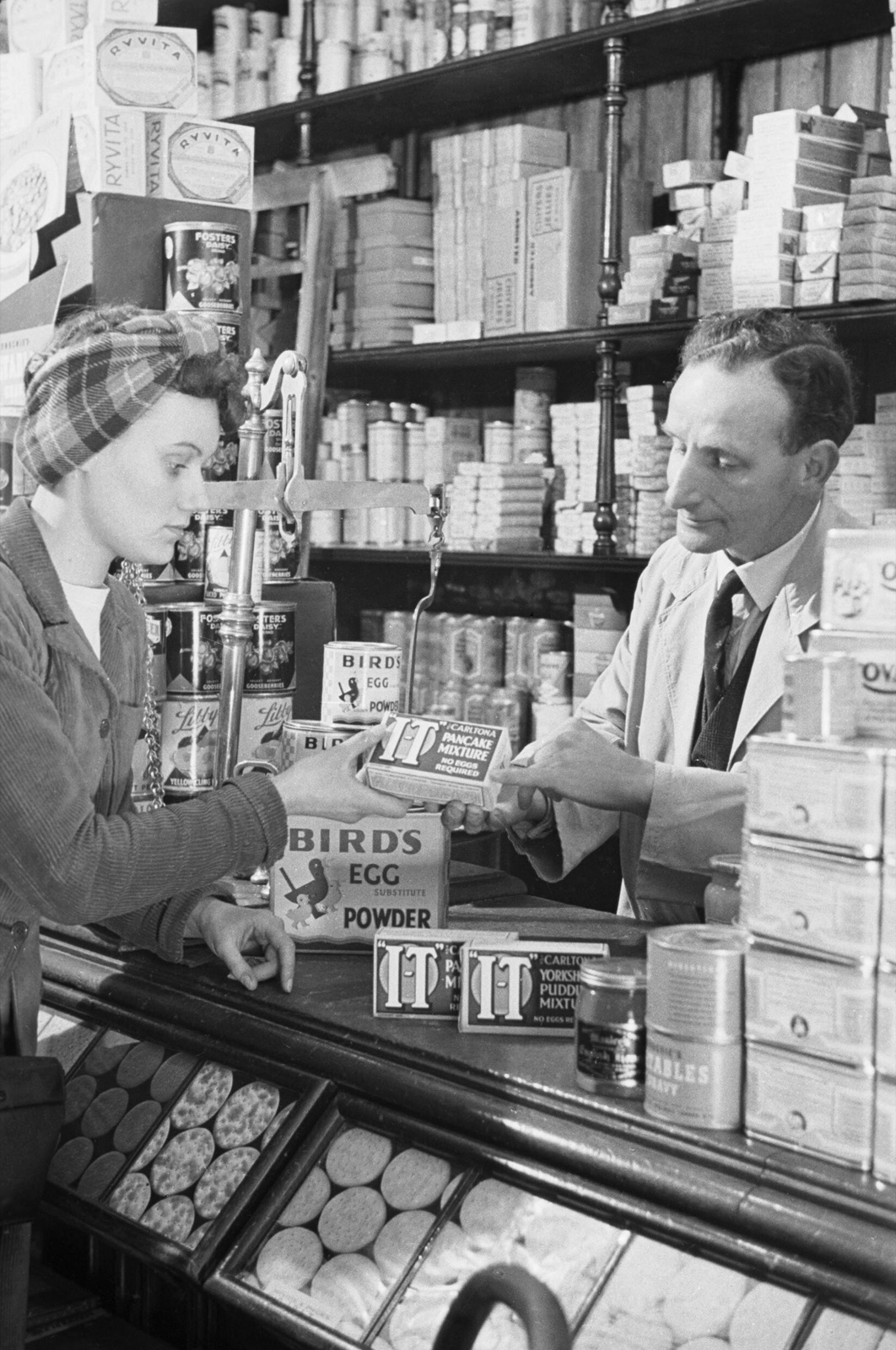 A customer is served across the counter of a small grocery shop, with tins and packaged goods stacked on the shelves behind the shopkeeper; a black-and-white photograph from Second World War Britain.
